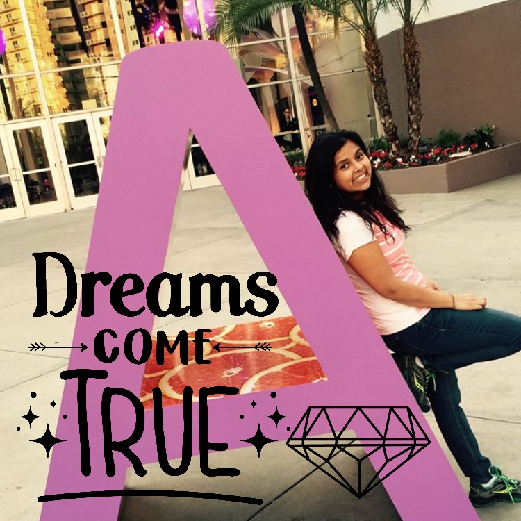 Smiling woman seated beside a large lavender letter installation in a bright outdoor setting.